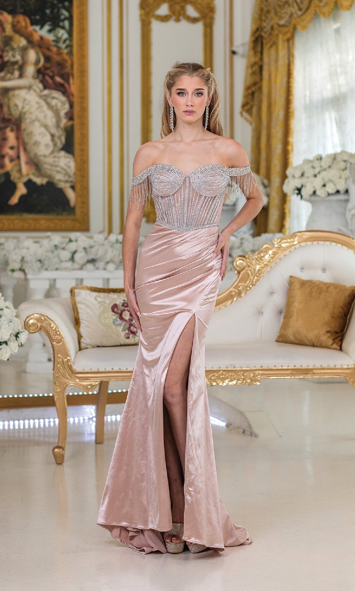 A woman poses in an ornate room wearing the Dancing Queen 4513 long prom dress, featuring a blush hue, high-leg slit, and sheer embellished bodice. She stands by a white and gold sofa amid elegant floral arrangements.